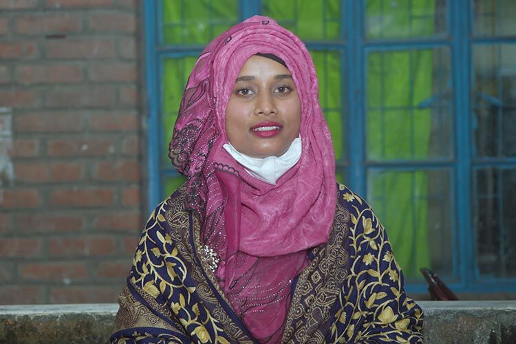 Woman looking to camera, with mask down, hands folded.