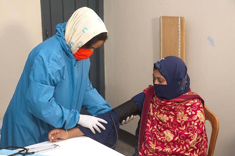 Woman in mask and protective gear measuring the blood pressure of another woman in mask sitting in chair. They are both in an office.